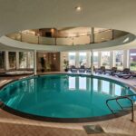 Innenpool mit runder Form, umgeben von Liegestühlen und großen Fensterfronten, mit Whirlpool im linken Bildhintergrund.